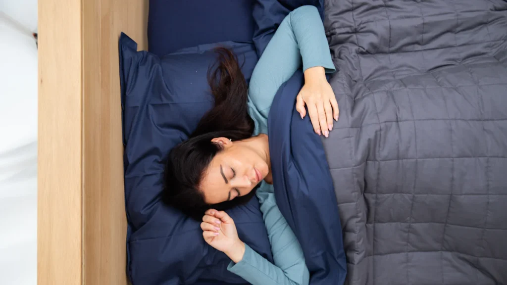 A person sleeps on a bed under a gray blanket, wearing a blue long-sleeve shirt, next to a wooden bedside.