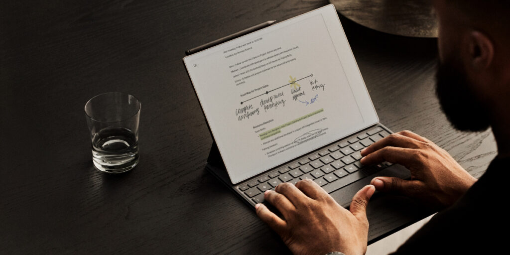 A person types on a laptop with a document on the screen. A glass of water is placed beside the laptop on the dark surface.