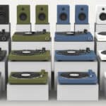 Display of various turntables and speakers in multiple colors including black, green, blue, and white, arranged on a white background.