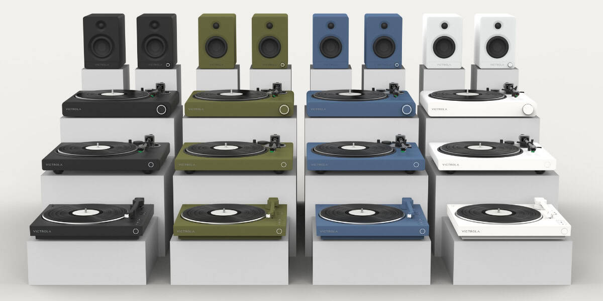 Display of various turntables and speakers in multiple colors including black, green, blue, and white, arranged on a white background.