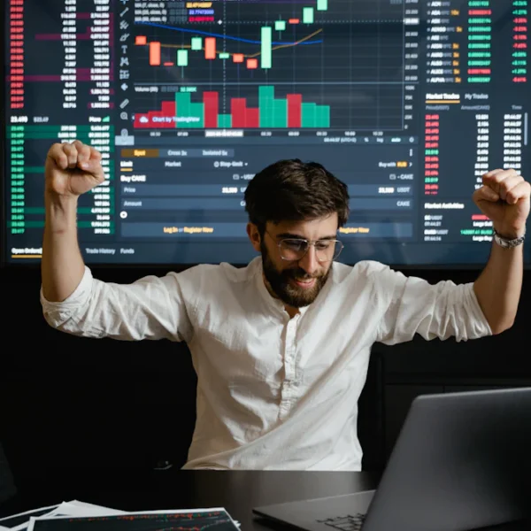 A person in a white shirt raises their fists in celebration in front of a laptop, with a large screen showing financial graphs and charts in the background.