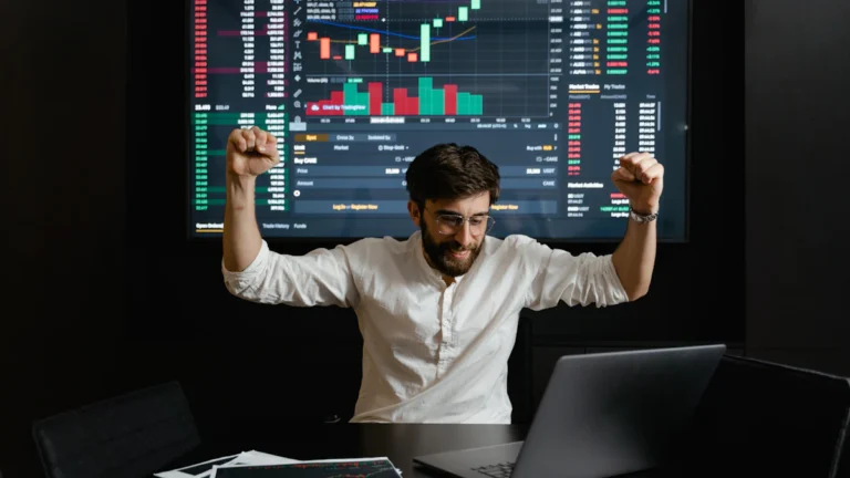 A person in a white shirt raises their fists in celebration in front of a laptop, with a large screen showing financial graphs and charts in the background.