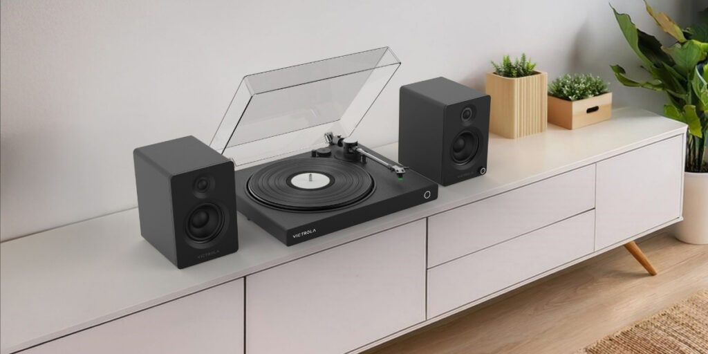 A record player with open dust cover, flanked by two speakers, sits on a white sideboard. A plant is on the right, and a small potted plant is on the left.