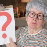 An older woman with gray hair and glasses holds up a clipboard displaying a large red question mark, sitting indoors on a couch.