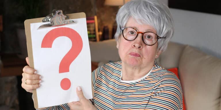 An older woman with gray hair and glasses holds up a clipboard displaying a large red question mark, sitting indoors on a couch.