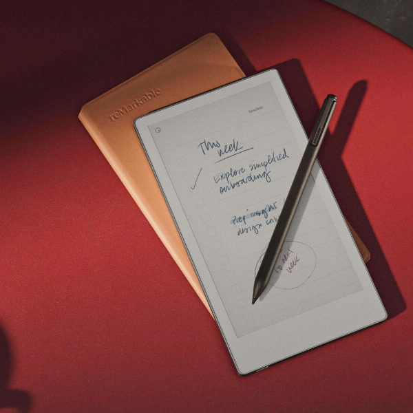 A digital notepad with handwritten notes and a stylus rests on a red surface beside a closed brown notebook. Part of a coffee cup is visible in the corner.