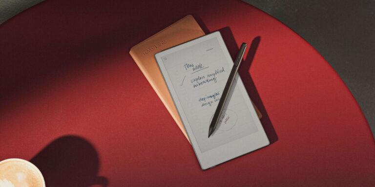 A digital notepad with handwritten notes and a stylus rests on a red surface beside a closed brown notebook. Part of a coffee cup is visible in the corner.
