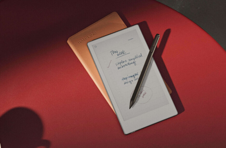 A digital notepad with handwritten notes and a stylus rests on a red surface beside a closed brown notebook. Part of a coffee cup is visible in the corner.