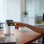 A person sits at a desk using a smartphone to make a contactless payment on a small terminal next to a laptop in an office setting.