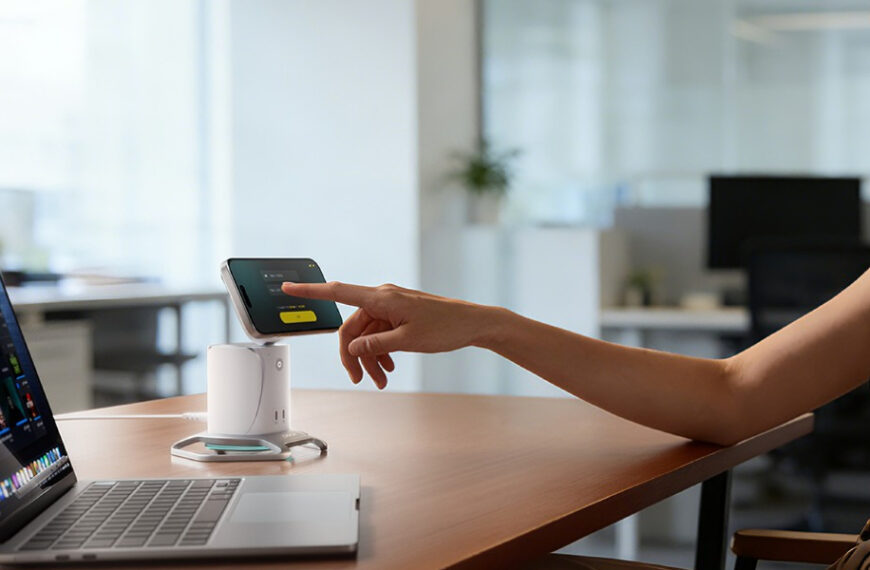 A person sits at a desk using a smartphone to make a contactless payment on a small terminal next to a laptop in an office setting.