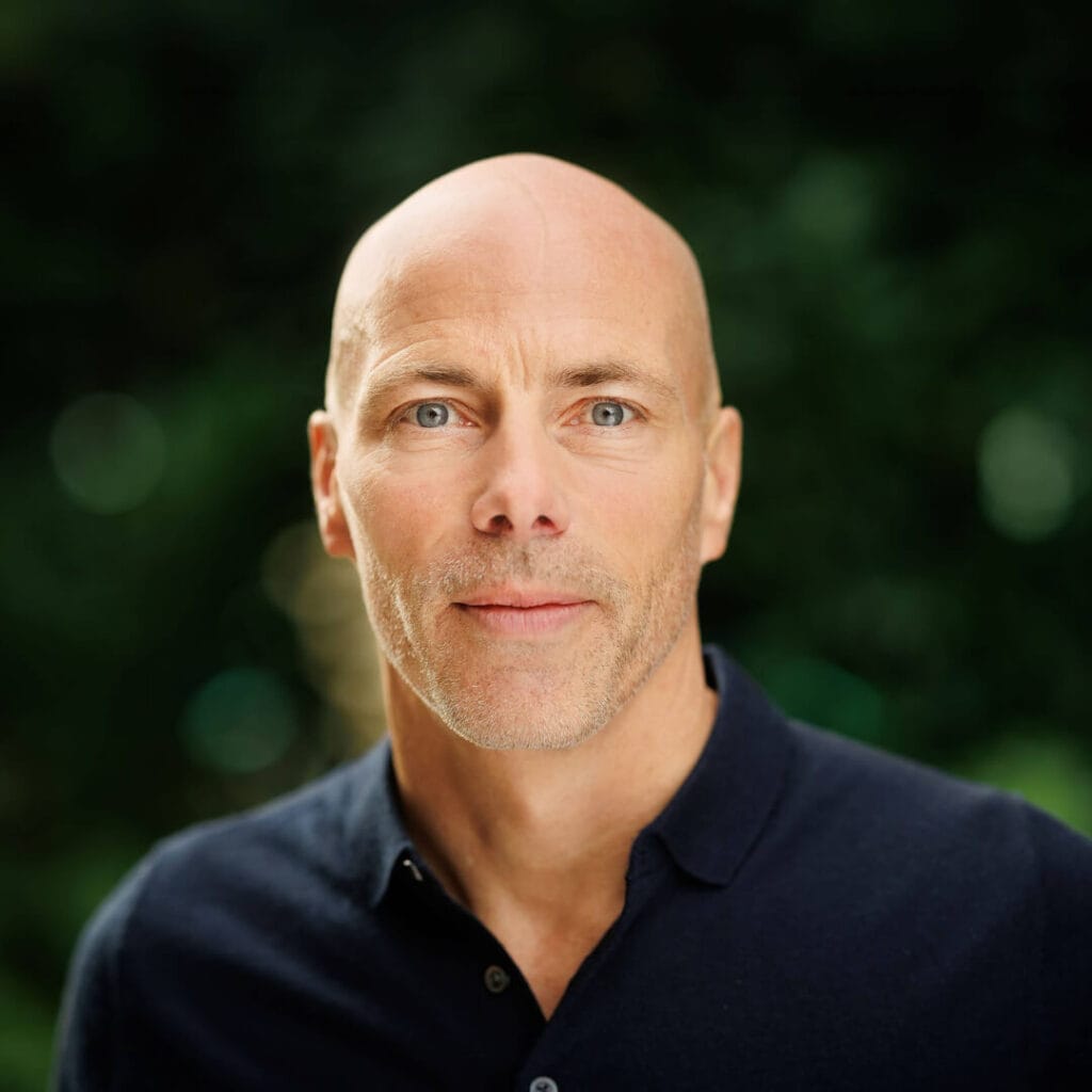 A middle-aged man with a shaven head and light facial hair, wearing a dark collared shirt, is looking at the camera against a blurred green background.