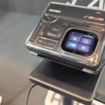 A compact digital camera labelled “Aurzen J’ZIP Pro” is displayed on a stand, showing a small touchscreen interface with various icons.