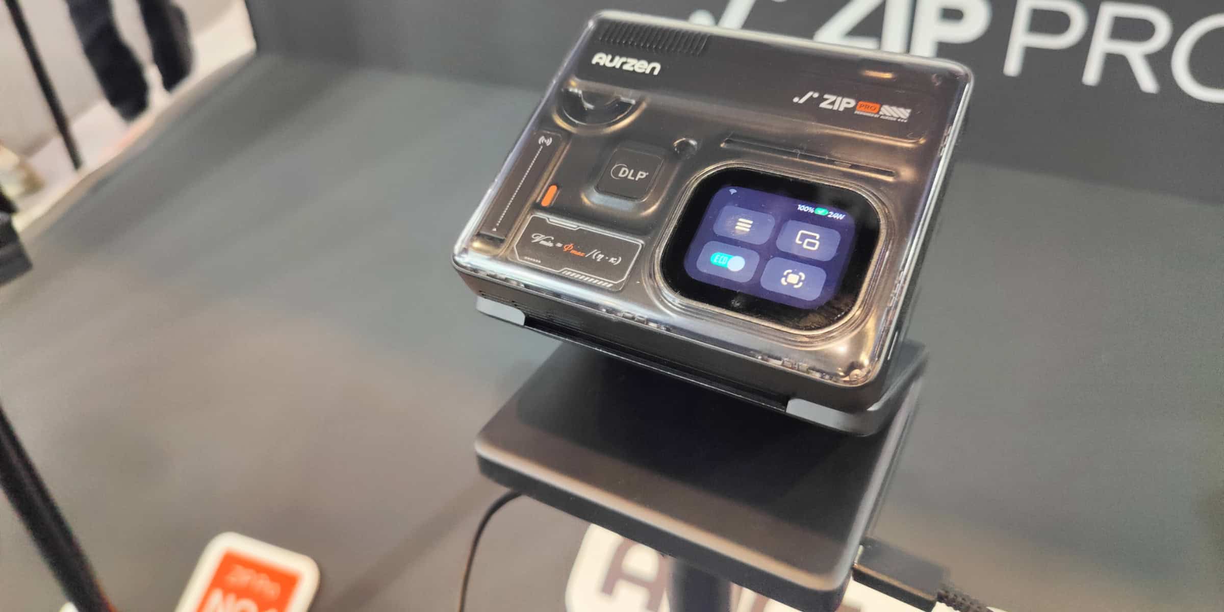 A compact digital camera labelled “Aurzen J’ZIP Pro” is displayed on a stand, showing a small touchscreen interface with various icons.