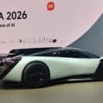 A futuristic white and black sports car is displayed at an indoor event with a large sign reading “Xiaomi IFA 2026 The Surging Wave of AI” in the background.